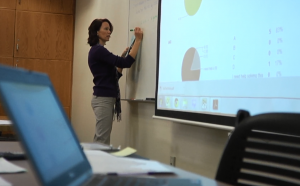 Female professor writing on a whiteboard, next to a screen projection of instructional materials for a math class.